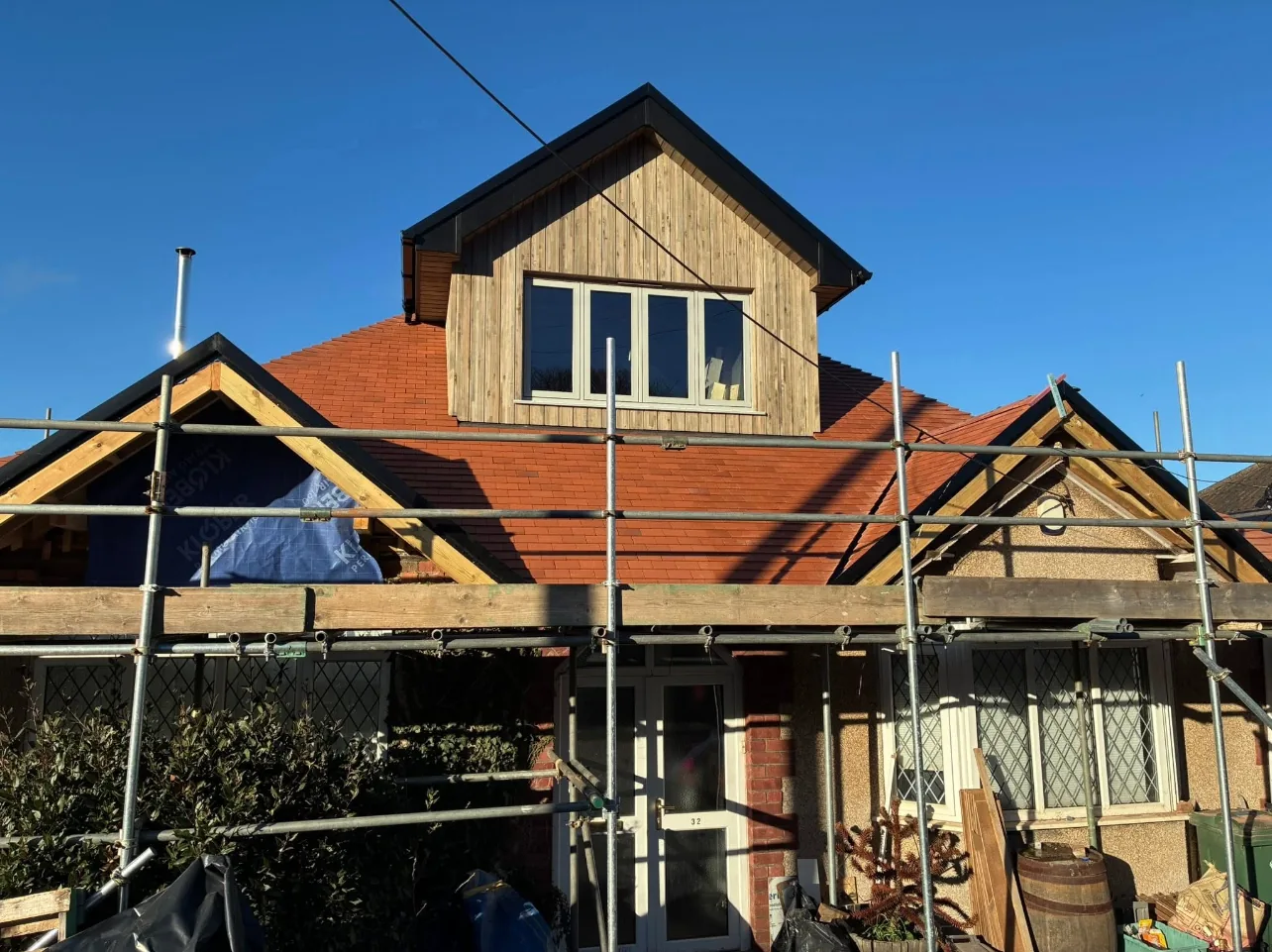 A timber-clad dormer with a four-pane window above a new clay-tiled roof on a bungalow, scaffolded, under a clear blue sky