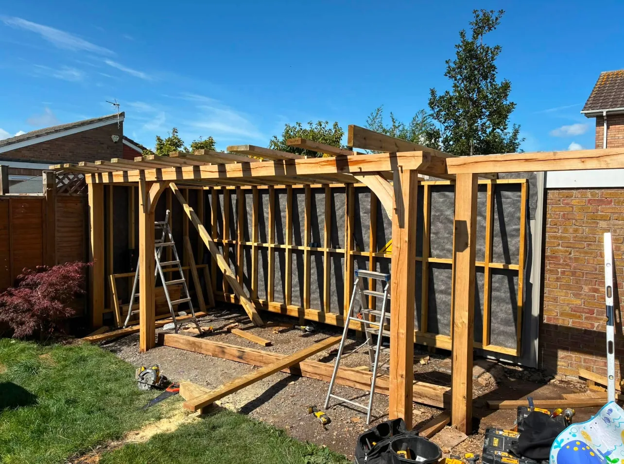 An oak-framed garden structure under construction, with curved braces, a rafter frame and timber studwork