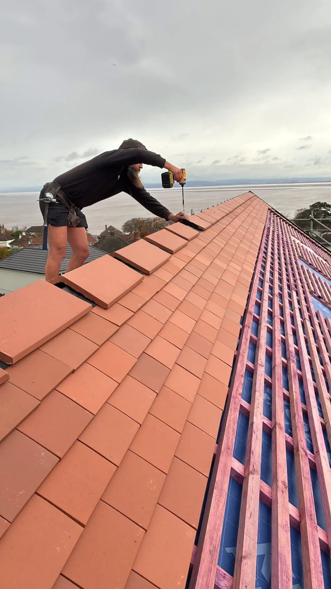 A roofer fixing ridge tiles on a newly tiled clay roof, with battens and membrane still open on the far slope and water beyond